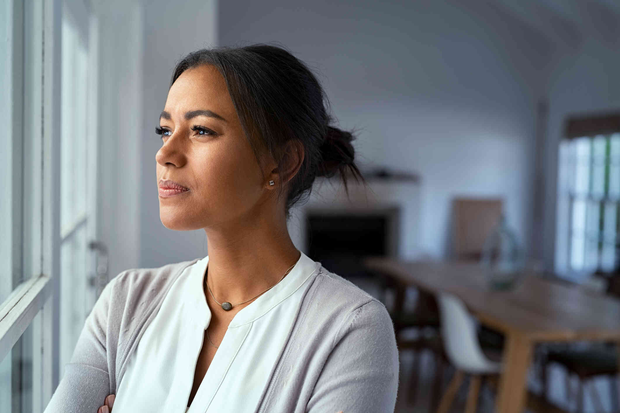 A woman in a white shrt stands in her home and gazes off deep in thought.