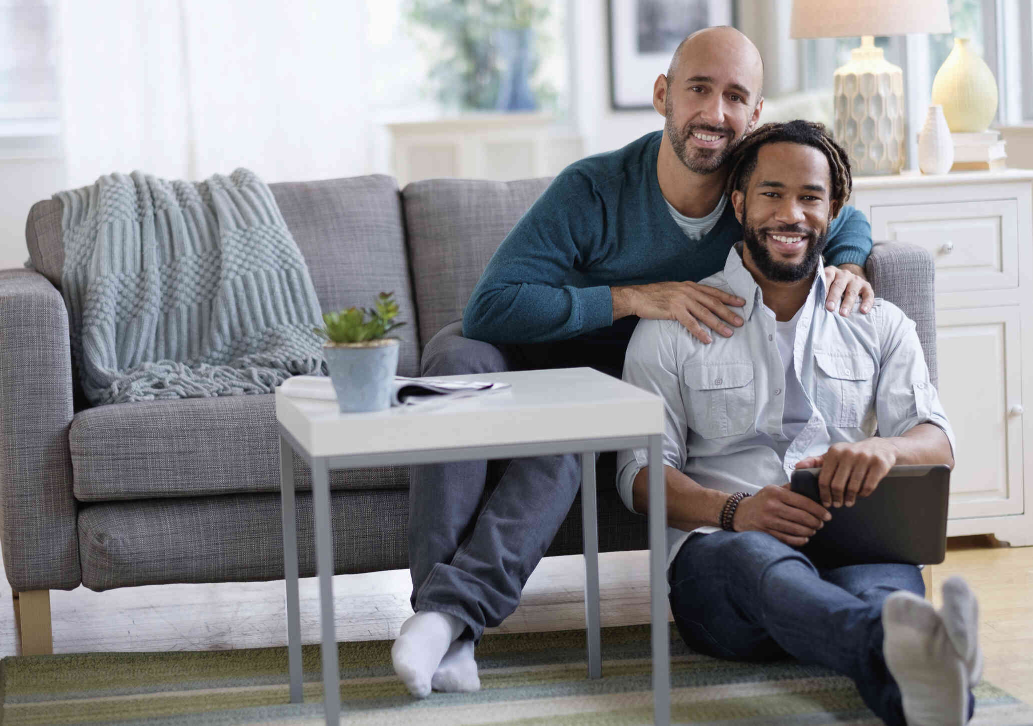 A man in a blue sweater sits on the couch while his male partner sits on the floor infront of him with a tablet as they both smile at the camera.