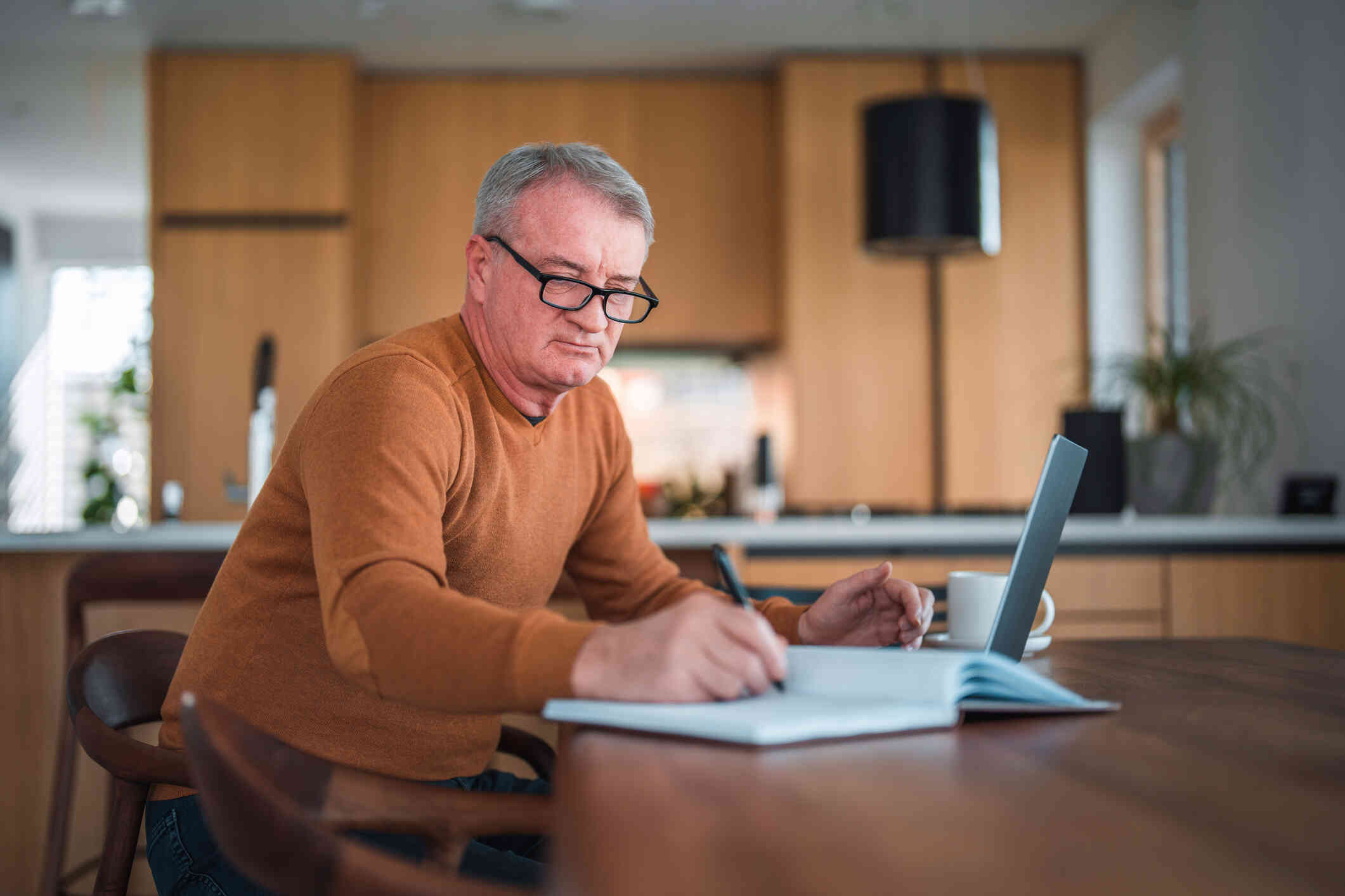 A middle aged man in an ornage shirt sits at his counter and takes notes in a notebook with his laptop open infront of him.