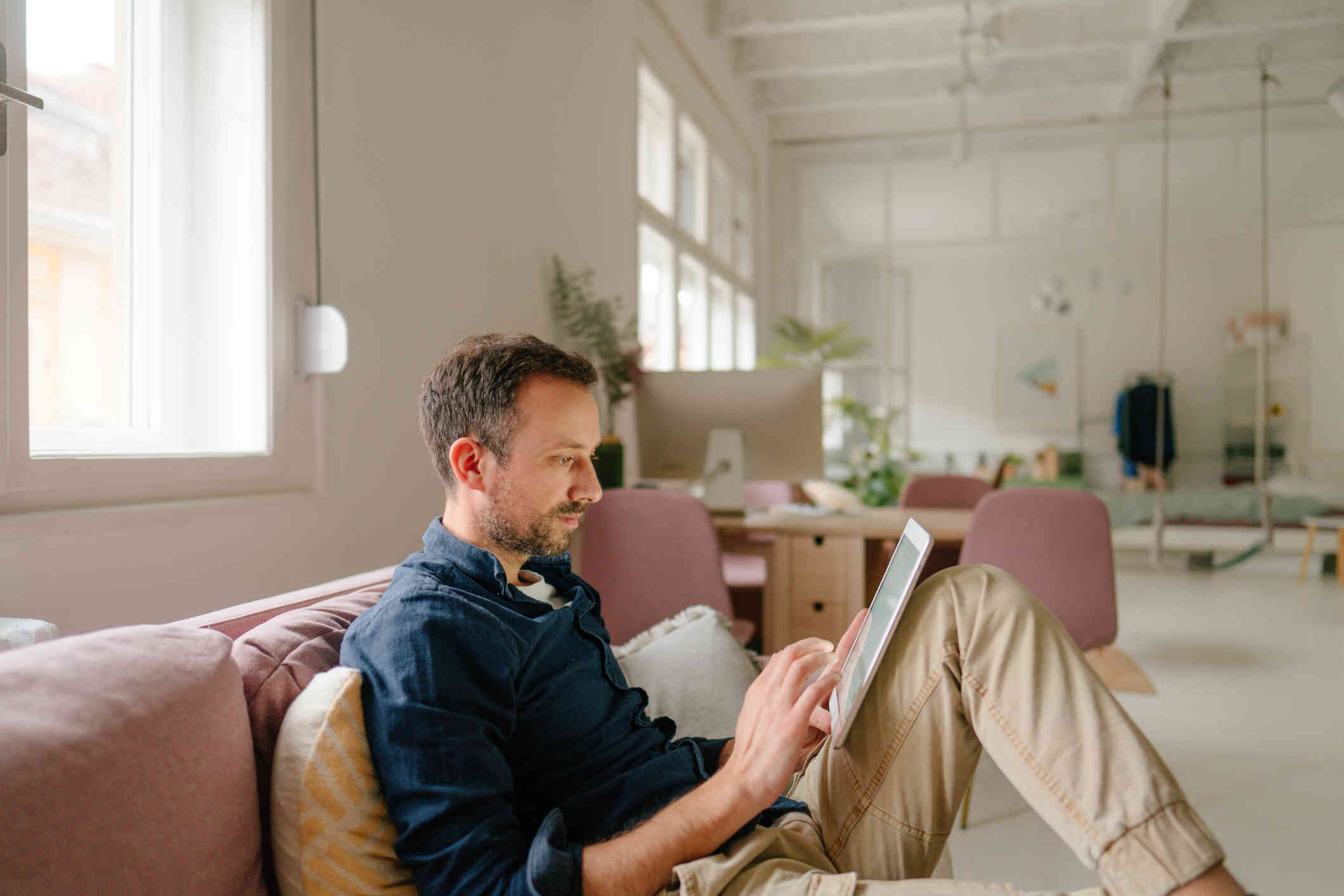 A man ina blue shirt sits on a pink couch in his home and looks at the tablet in his hand.